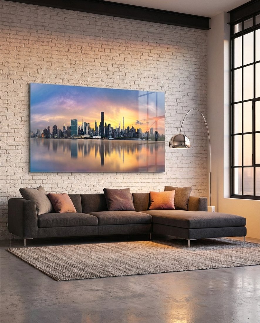 Manhattan skyline at sunset mirrored in the river, panoramic glass wall art above a sectional sofa by a brick wall