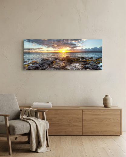 Rocky shore ocean sunset panoramic glass wall art above a light wood sideboard beside a gray armchair
