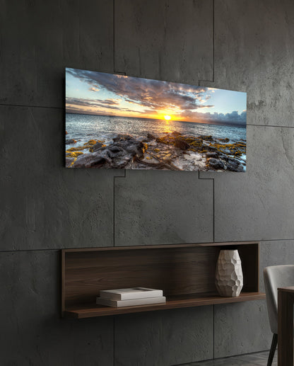 Rocky shore ocean sunset panoramic glass wall art above a floating wood shelf on a dark stone wall