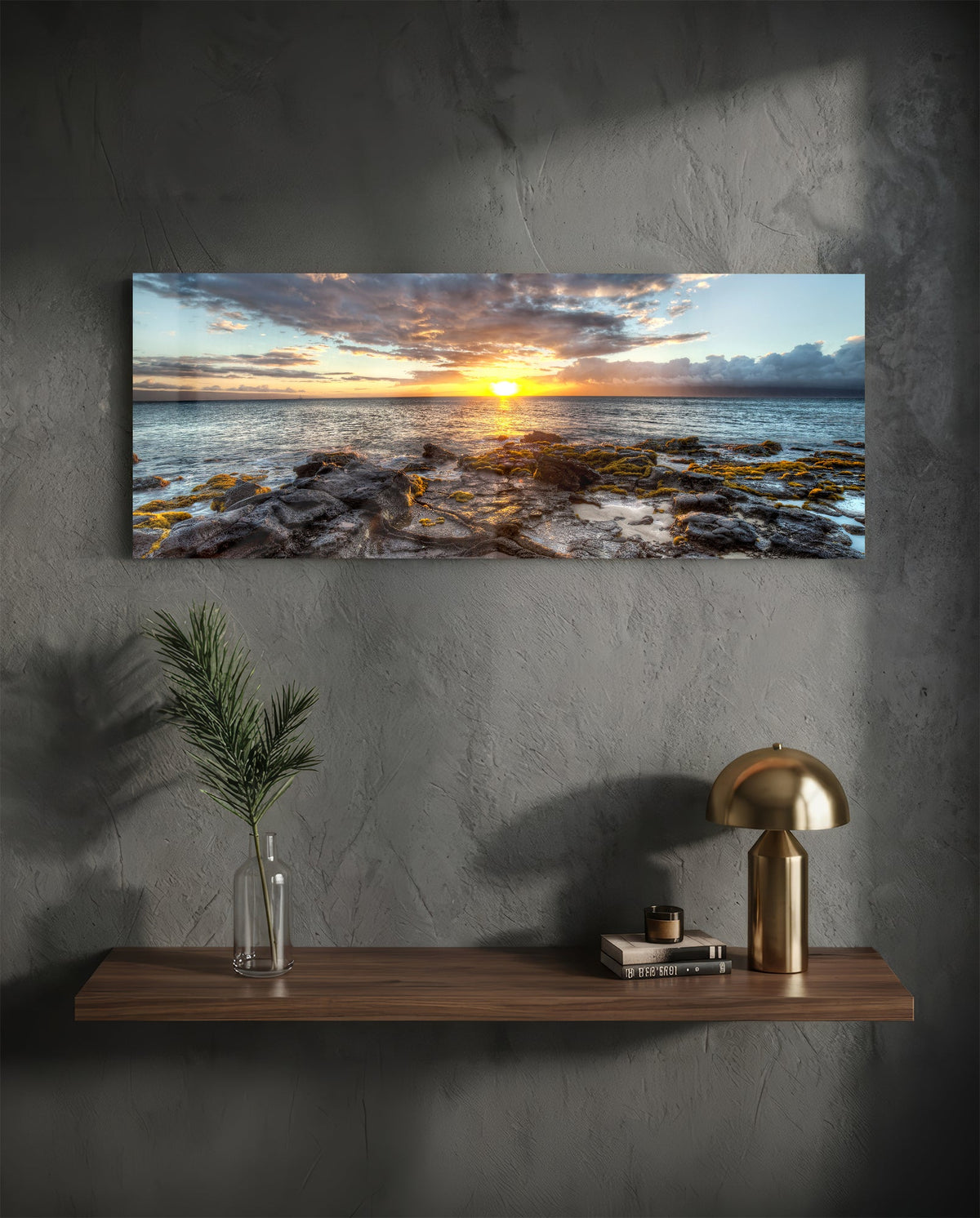 Rocky shore ocean sunset panoramic glass wall art above a wood shelf with a brass lamp on a dark wall
