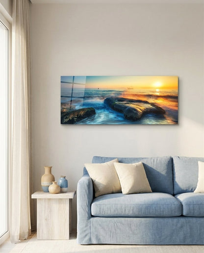 Mossy rocks sunrise sea panoramic glass wall art above a blue sofa on a beige wall