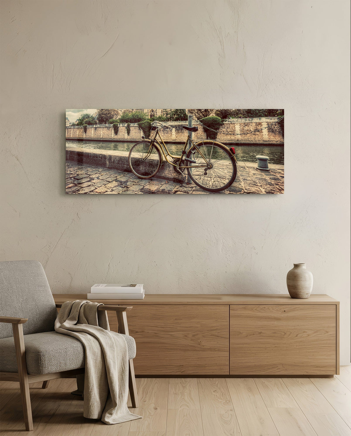 Vintage riverside bicycle glass wall art above a light wood sideboard beside a gray armchair with a throw
