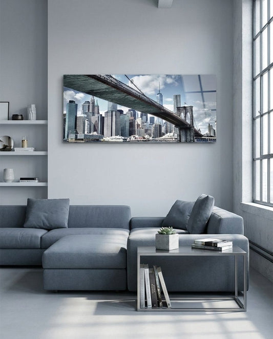 Brooklyn Bridge and Manhattan skyline panoramic glass wall art above a gray sectional sofa by tall windows