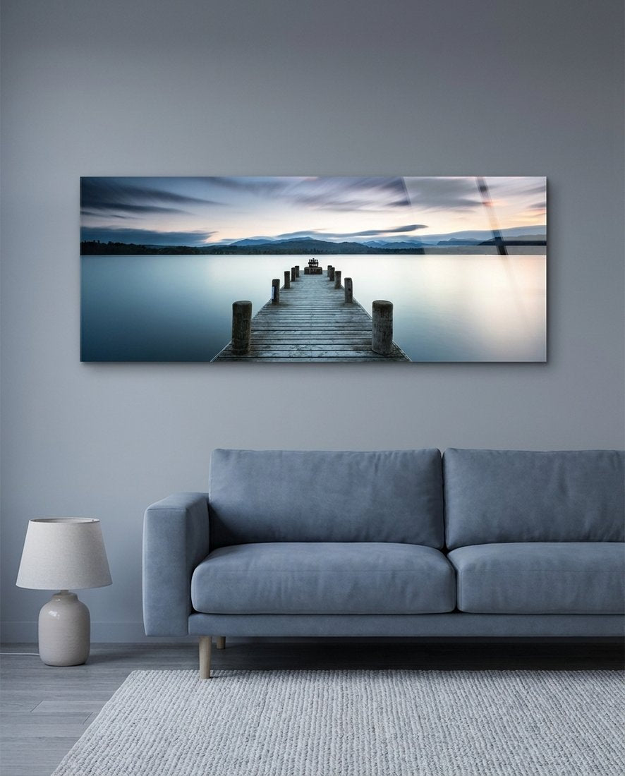 Blue hour lake jetty panoramic glass wall art above a blue sofa on a gray-blue wall