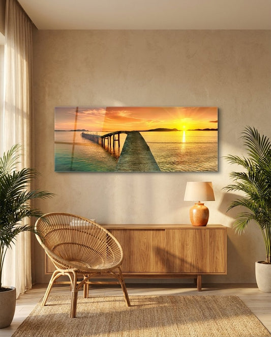 Golden sunset curved pier panoramic glass wall art above a wooden sideboard beside a rattan chair and plants