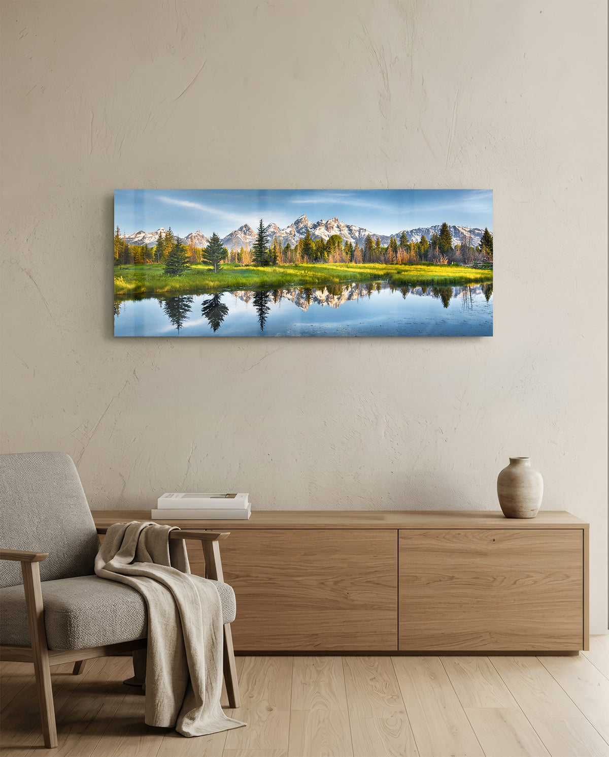Mountain lake reflection glass wall art above a light wood sideboard beside a gray armchair with a throw