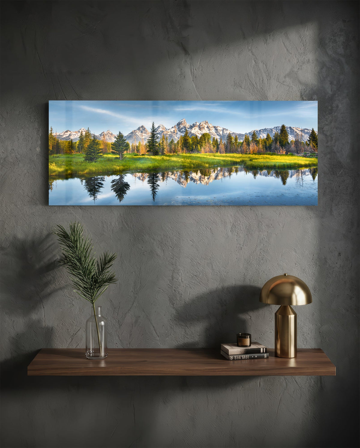 Mountain lake reflection glass wall art on a charcoal wall above a floating shelf with a brass lamp and greenery