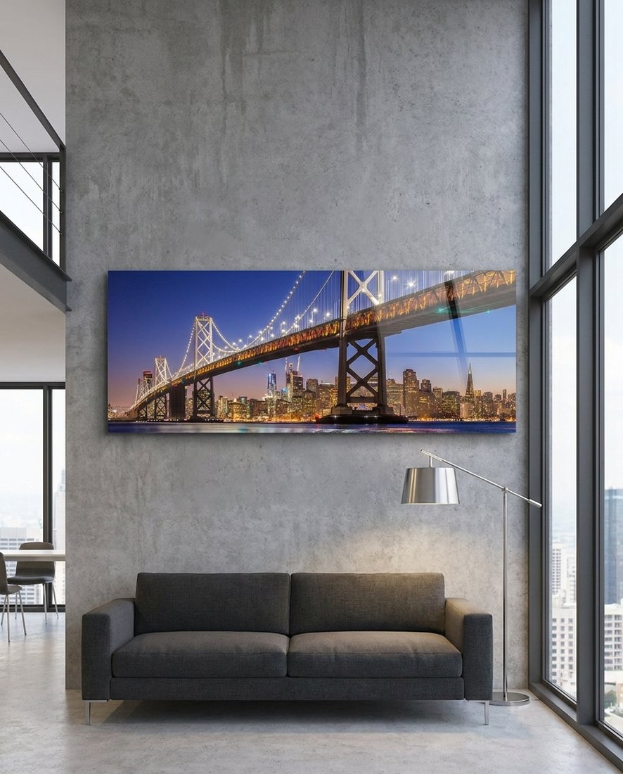 San Francisco Bay Bridge lit at dusk over the city skyline, panoramic glass wall art above a dark sofa in a loft