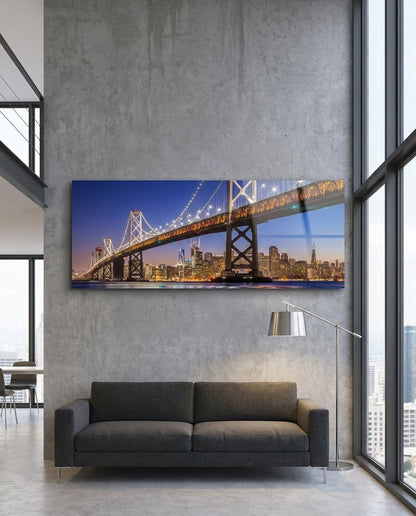 San Francisco Bay Bridge lit at dusk over the city skyline, panoramic glass wall art above a dark sofa in a loft