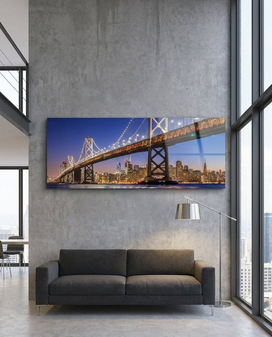 San Francisco Bay Bridge lit at dusk over the city skyline, panoramic glass wall art above a dark sofa in a loft