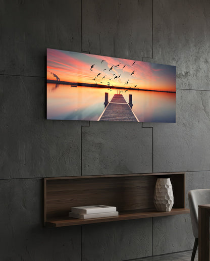 Sunset pier panoramic glass wall art on a dark stone wall above a walnut floating shelf