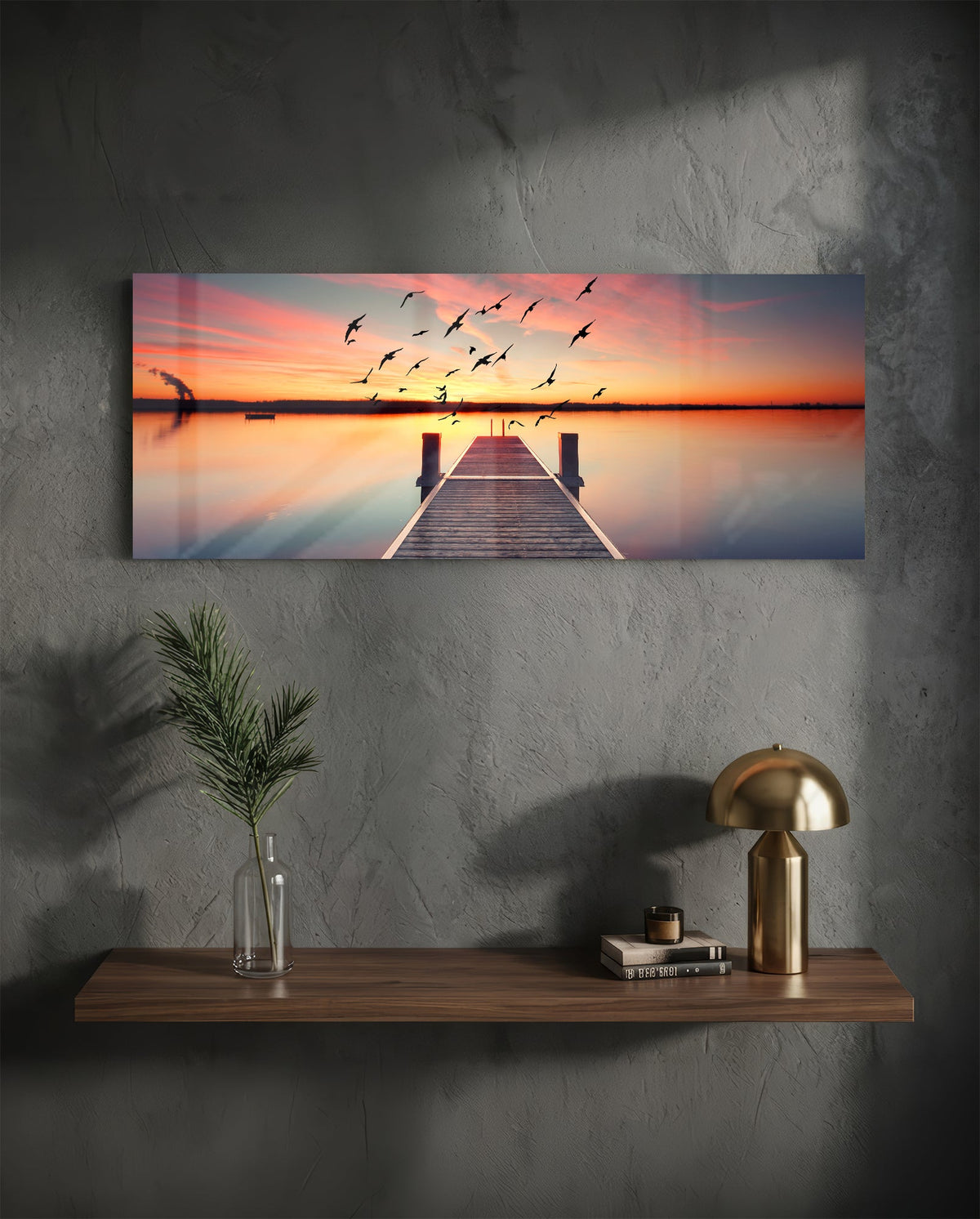 Sunset pier panoramic glass wall art on a dark plaster wall above a shelf with a gold lamp