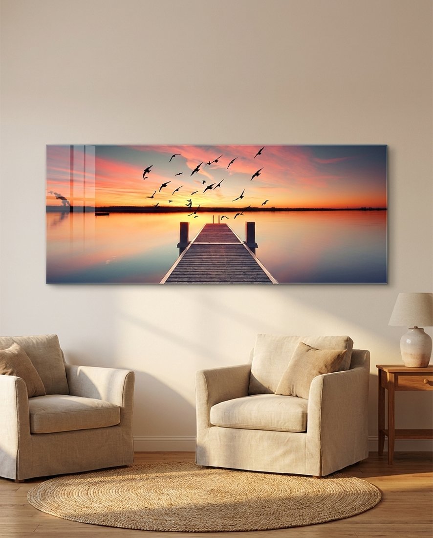 Wooden pier on still water under a pink and orange sunset with flying birds, panoramic glass wall art above armchairs
