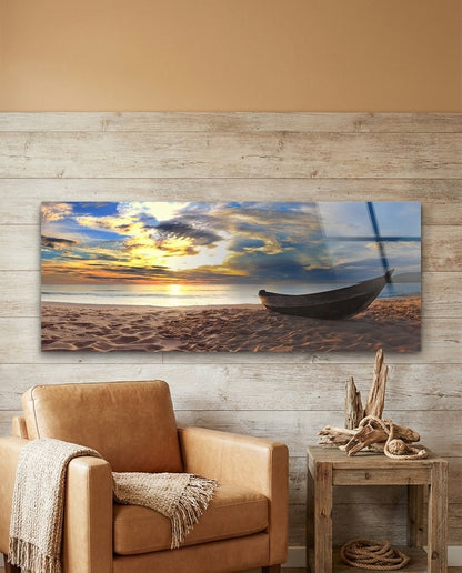 Wooden boat on a sandy beach at sunset panoramic glass wall art above a tan leather armchair on a rustic wood plank wall