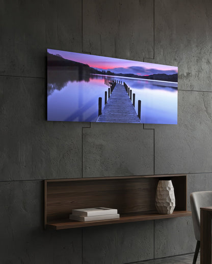 Purple twilight lake jetty glass wall art above a dark wood wall shelf on a charcoal paneled wall