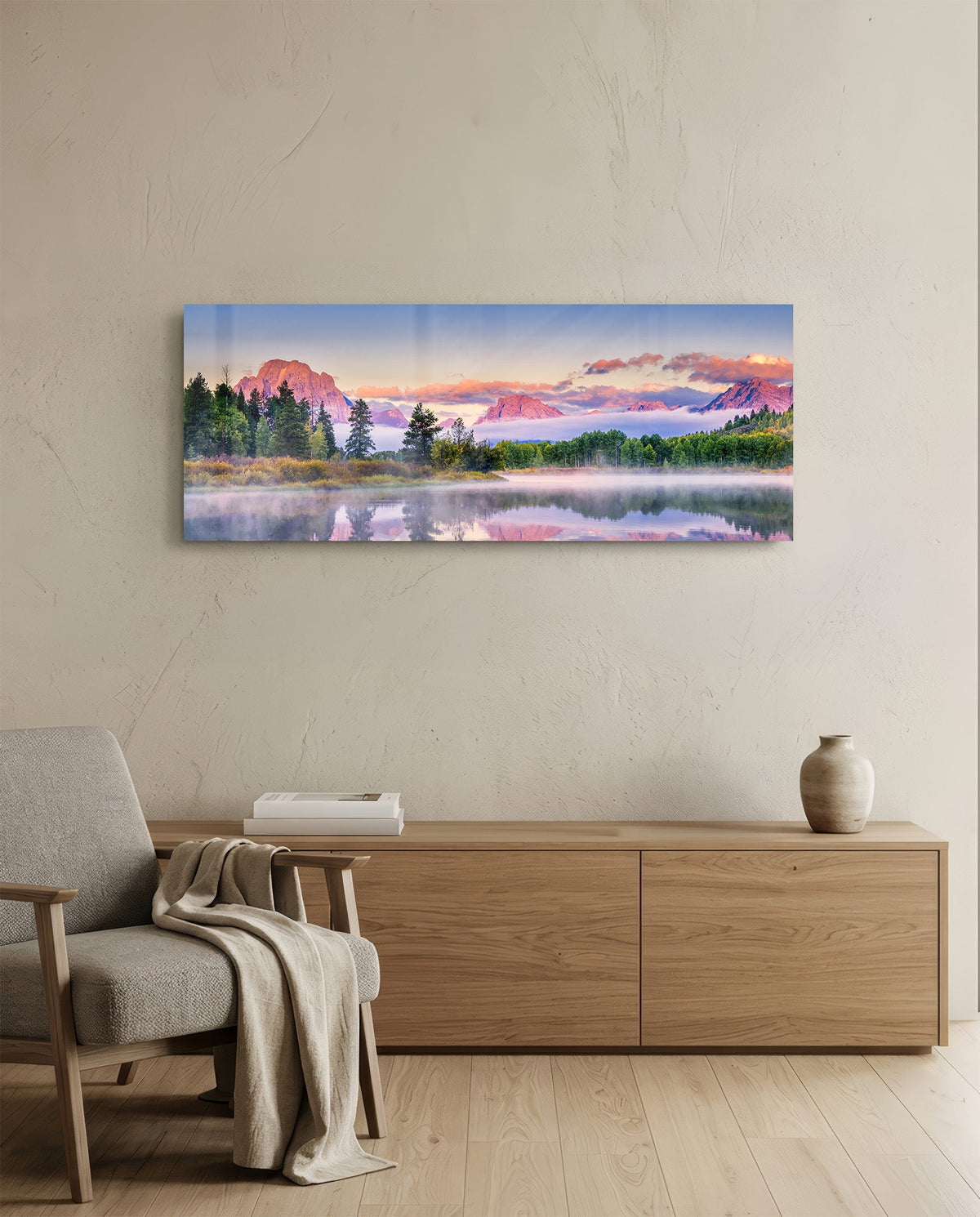 Pink dawn mountain lake panoramic glass wall art above a light wood sideboard beside a gray armchair