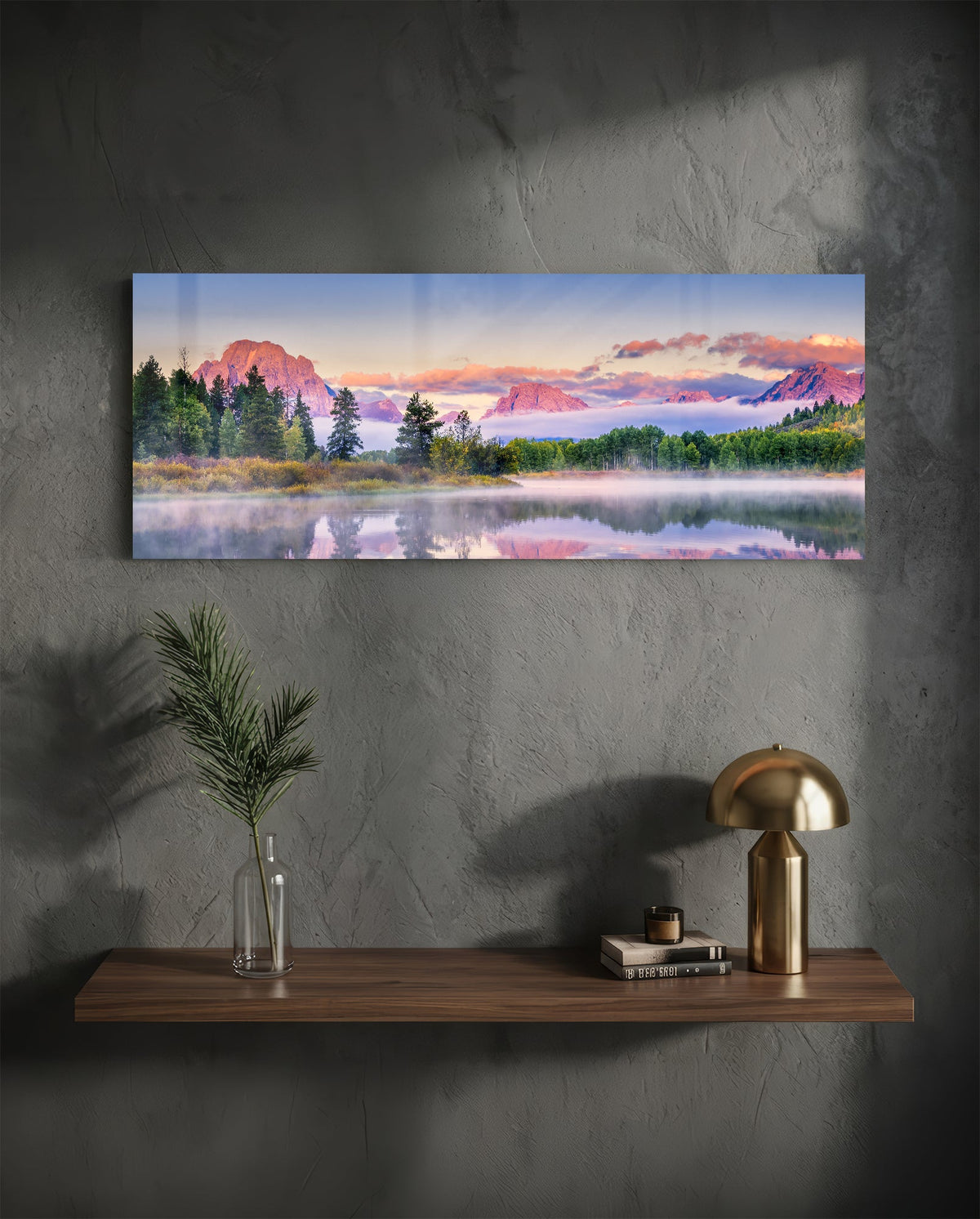 Pink dawn mountain lake panoramic glass wall art above a wood shelf with a brass lamp on a dark wall