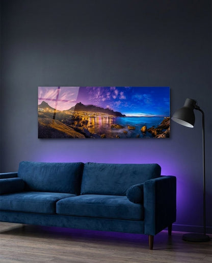 Seaside town lights under mountains at dusk, panoramic glass wall art above a blue velvet sofa with purple glow