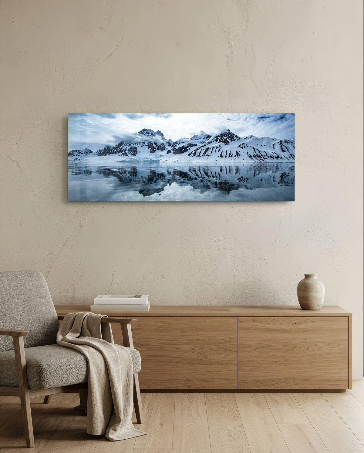 Snowy mountains icy lake panoramic glass wall art above a light wood sideboard beside a gray armchair