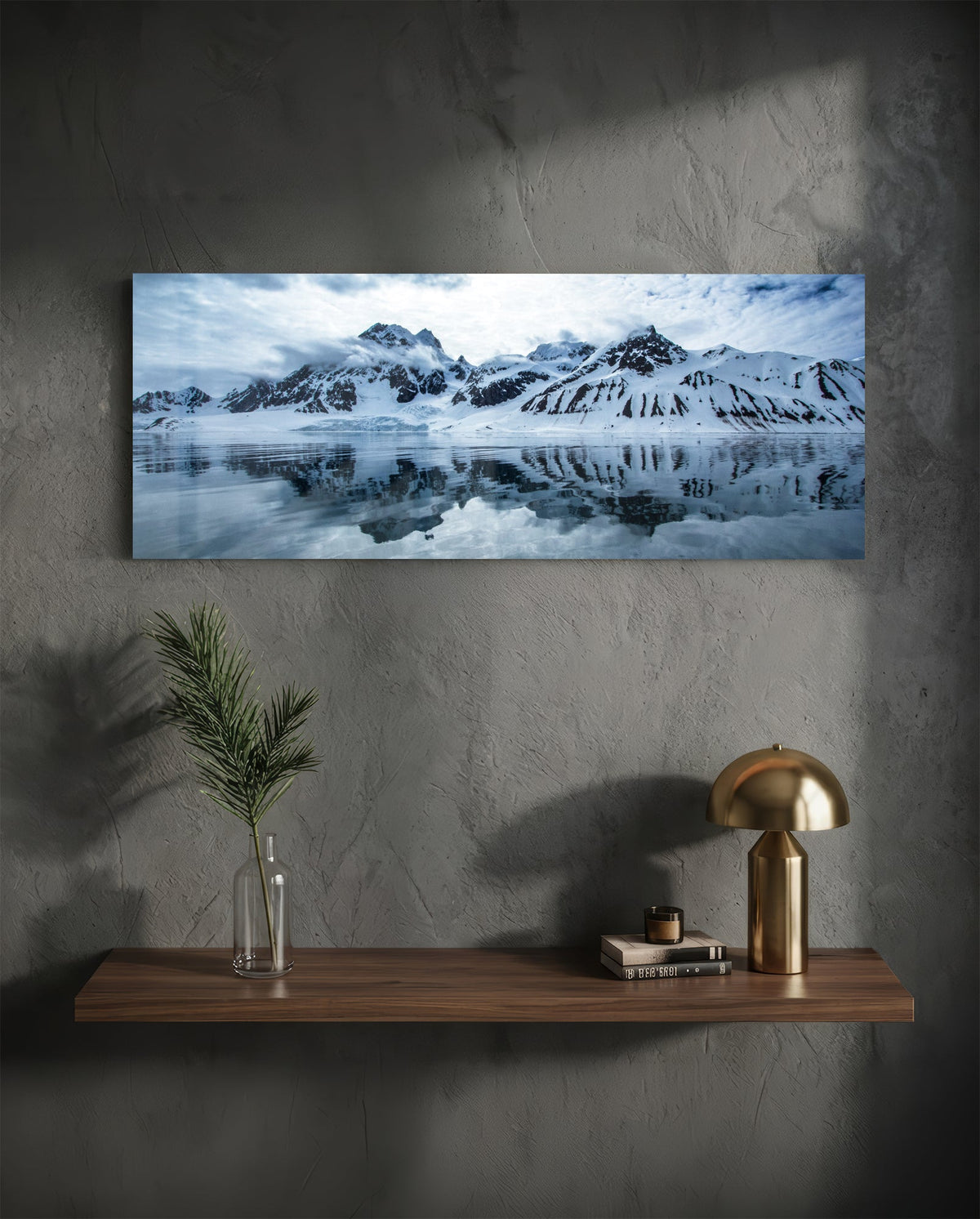 Snowy mountains icy lake panoramic glass wall art above a wood shelf with a brass lamp on a dark wall