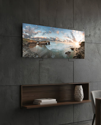 Rock arch coastline at sunset glass wall art above a dark wood wall shelf on a charcoal paneled wall