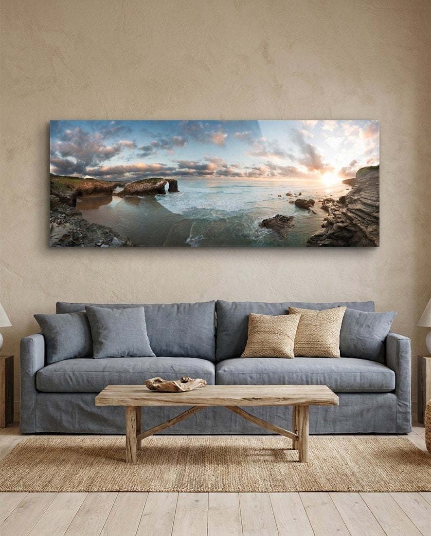 Rock arch coastline at sunset panoramic glass wall art above a gray sofa and a wooden coffee table