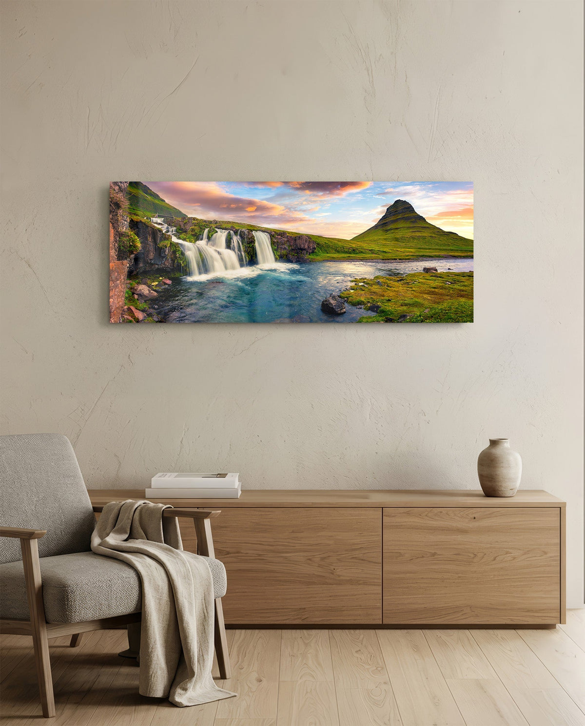 Waterfall and cone mountain panoramic glass wall art above a light wood sideboard beside a gray armchair