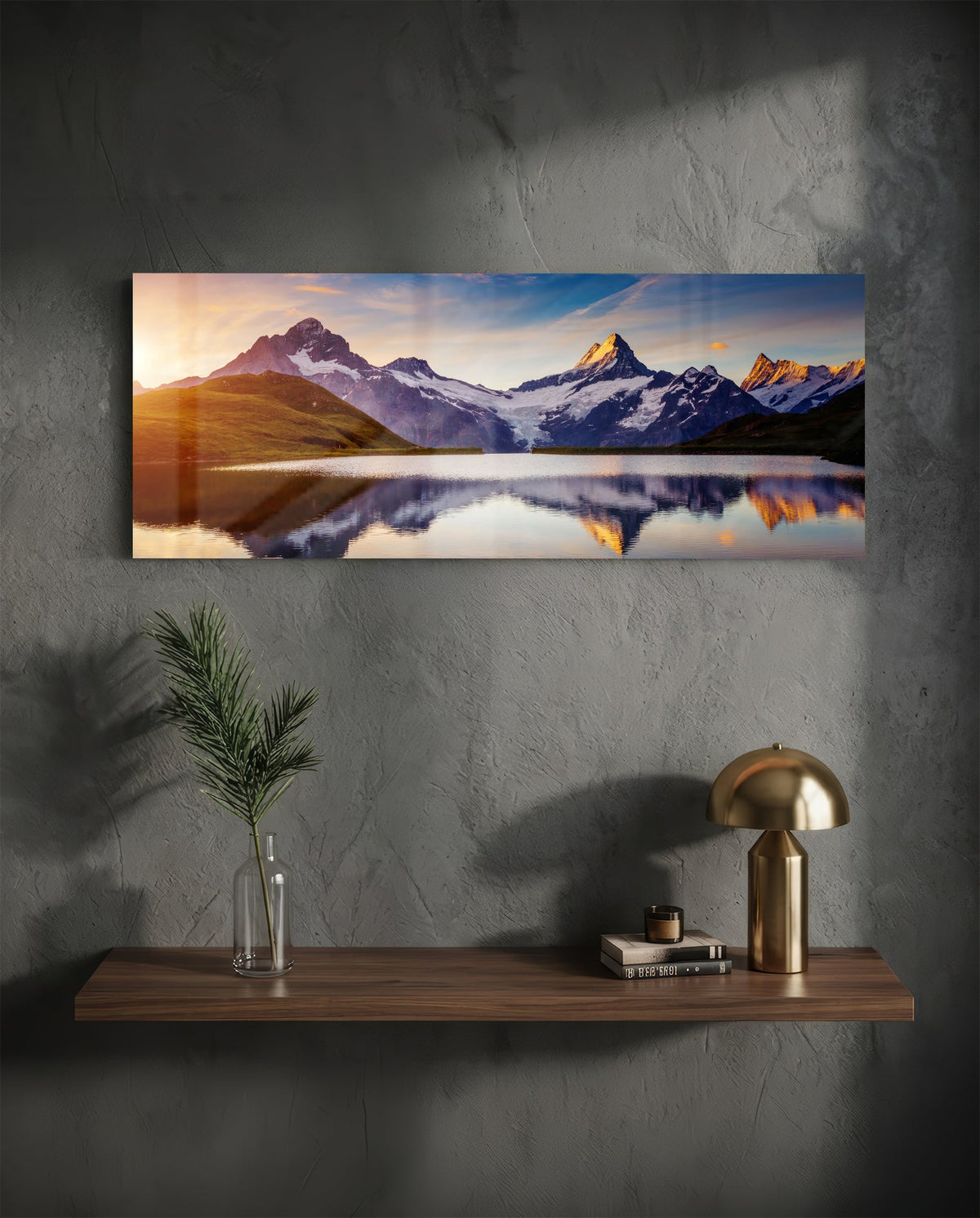 Sunrise alpine peaks lake panoramic glass wall art above a wood shelf with a brass lamp on a dark wall