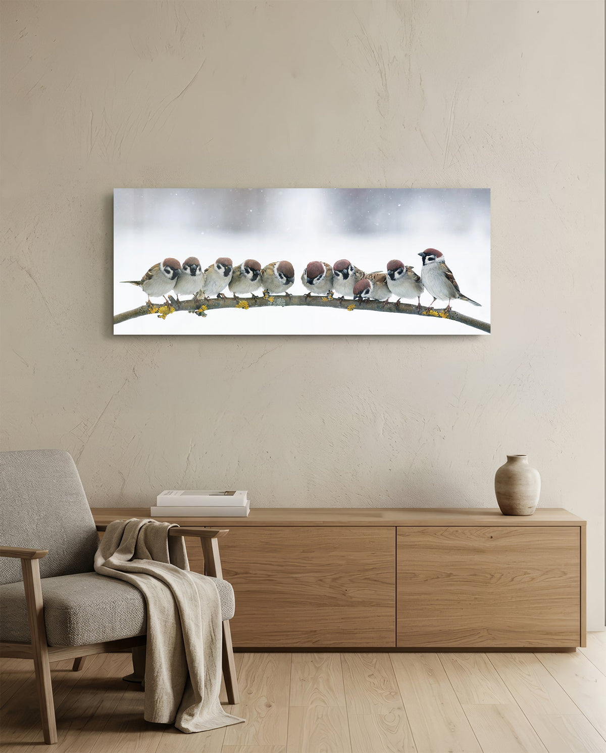 Sparrow flock in snow glass wall art above a low wood sideboard beside a gray armchair