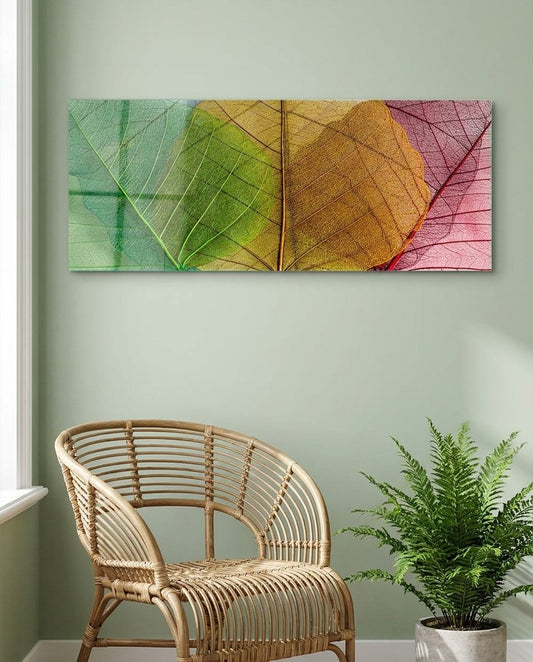 Veined leaves shading from green to orange and pink, panoramic glass wall art above a rattan chair and a fern