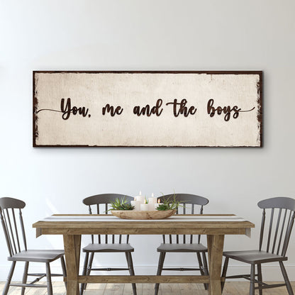 Long cream canvas sign with dark script reading You, me and the boys above a wood dining table and gray chairs