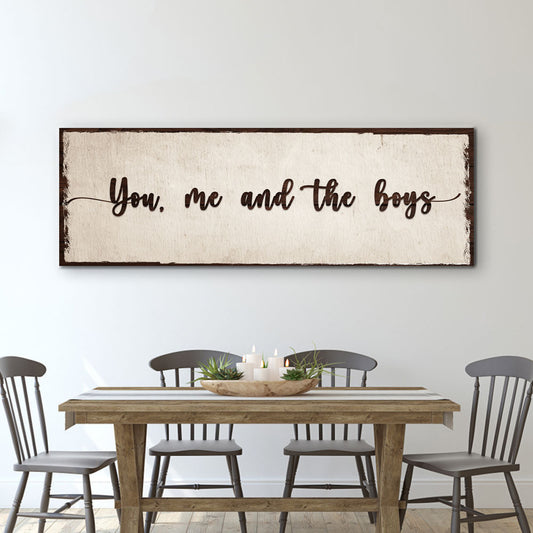 Long cream canvas sign with dark script reading You, me and the boys above a wood dining table and gray chairs