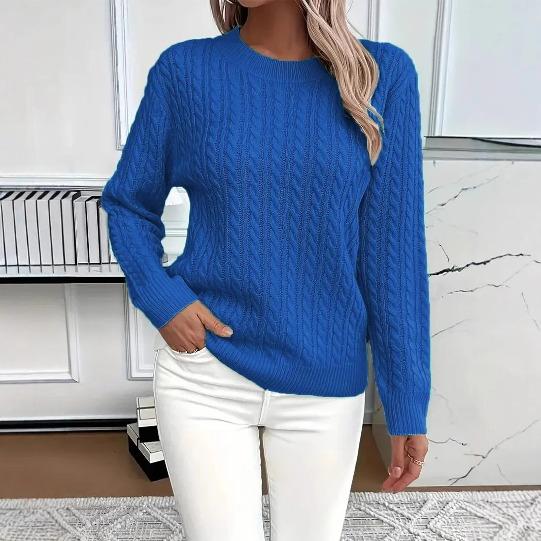 Blue women’s cable-knit sweater styled with white pants in a clean room setting