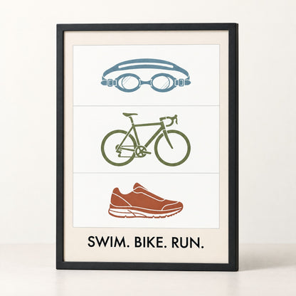 Framed swim bike run poster standing on a plain warm ground, black wood frame and matte print