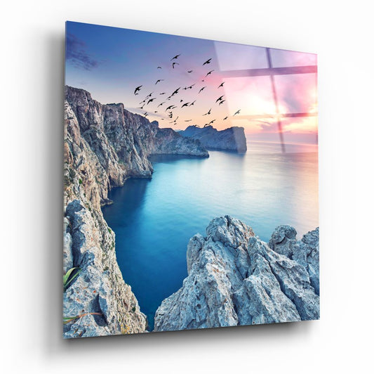 Pale rocky sea cliffs above a calm blue inlet with birds flying into a pink sunset, square glass wall art
