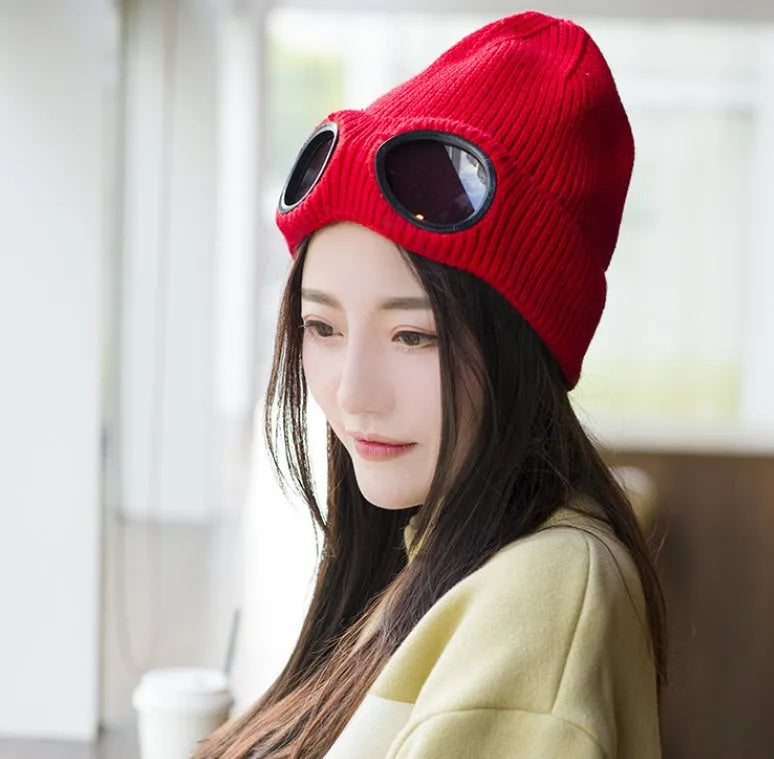 Model wearing red knitted wool hat with goggle lenses, indoor lifestyle view