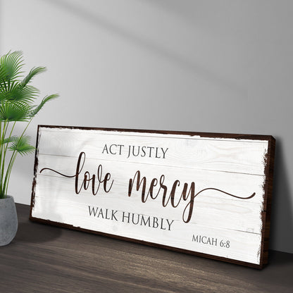 Angled view of the plank-look Act Justly Love Mercy canvas sign leaning on a dark wood floor