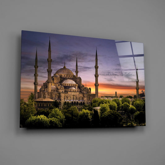 Mosque at purple dusk glass wall art hanging on a plain gray wall, seen at an angle