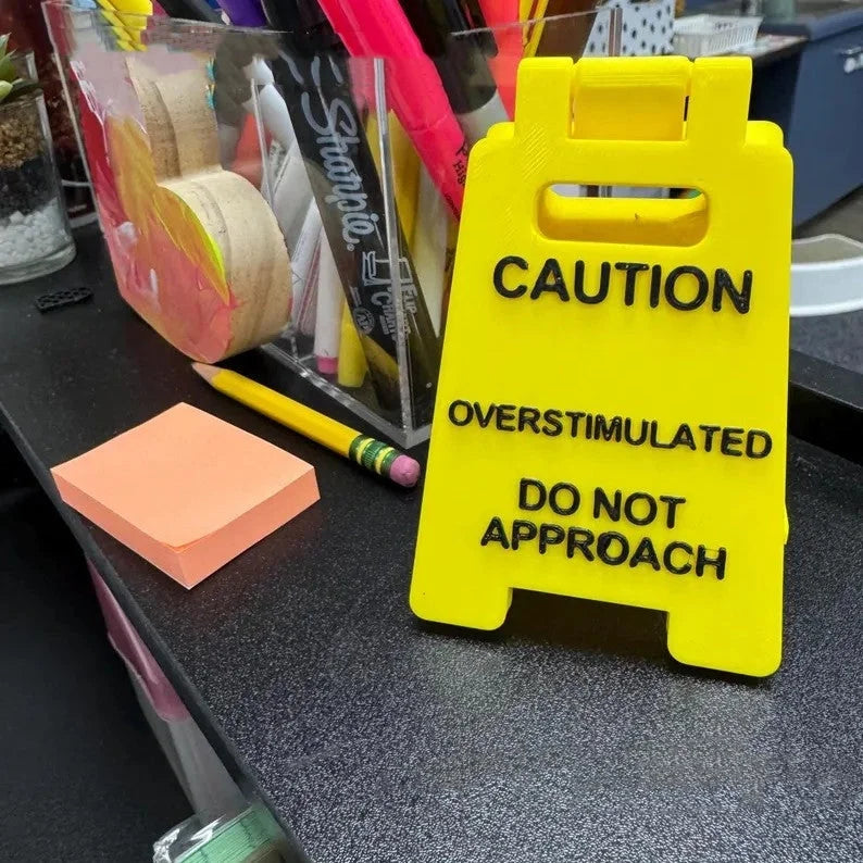Overstimulated caution sign placed near sticky notes and office supplies on a desktop.