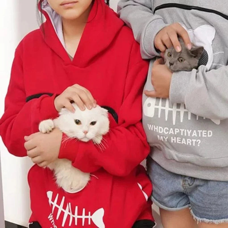 Person wearing a red pet-pouch hoodie while a white cat rests inside the front pocket.