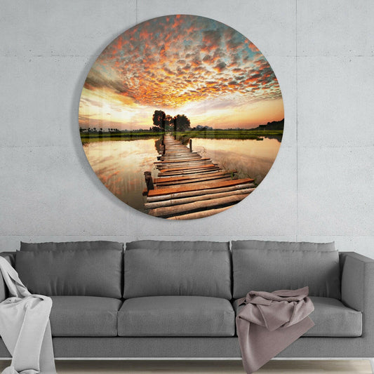 Wooden pier lake sunset round glass wall art above a gray sofa with white and mauve throws on a pale concrete wall