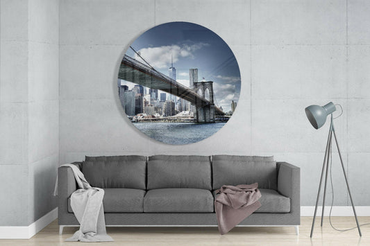 Brooklyn Bridge skyline round glass wall art above a gray sofa with draped throws on a pale concrete wall