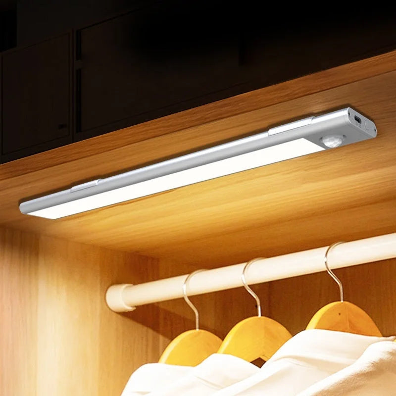 Silver rechargeable LED strip light installed in wooden wardrobe closet illuminating hanging clothes with warm light