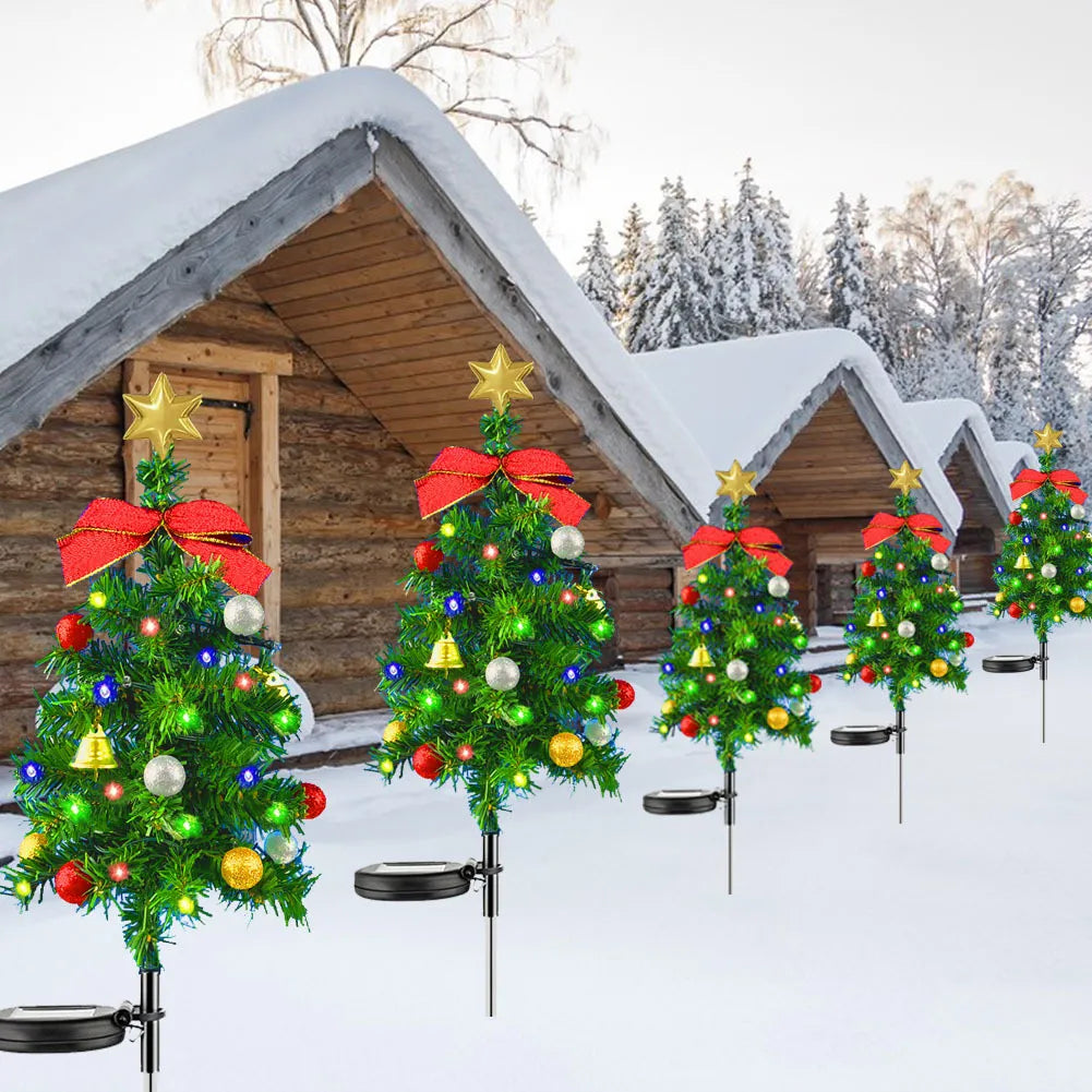 Small solar Christmas trees with lights and red bows in front of a wooden cabin in a snowy landscape.