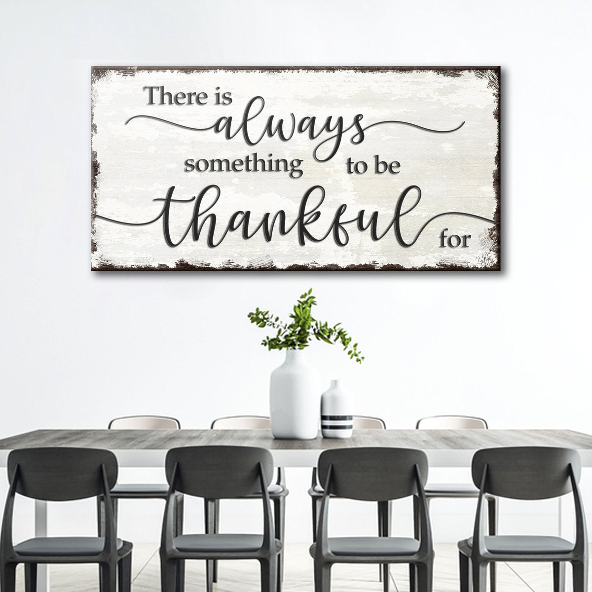 Wide white canvas sign with black script about being thankful and a dark worn edge above a dining table with black chairs