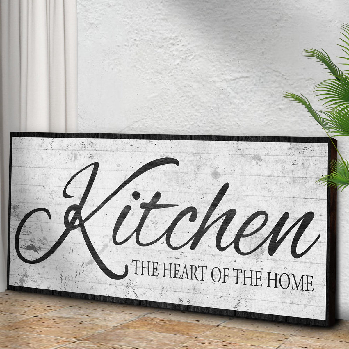 Kitchen Heart of the Home White Script canvas seen at an angle on a concrete wall above a wood floor, view 3