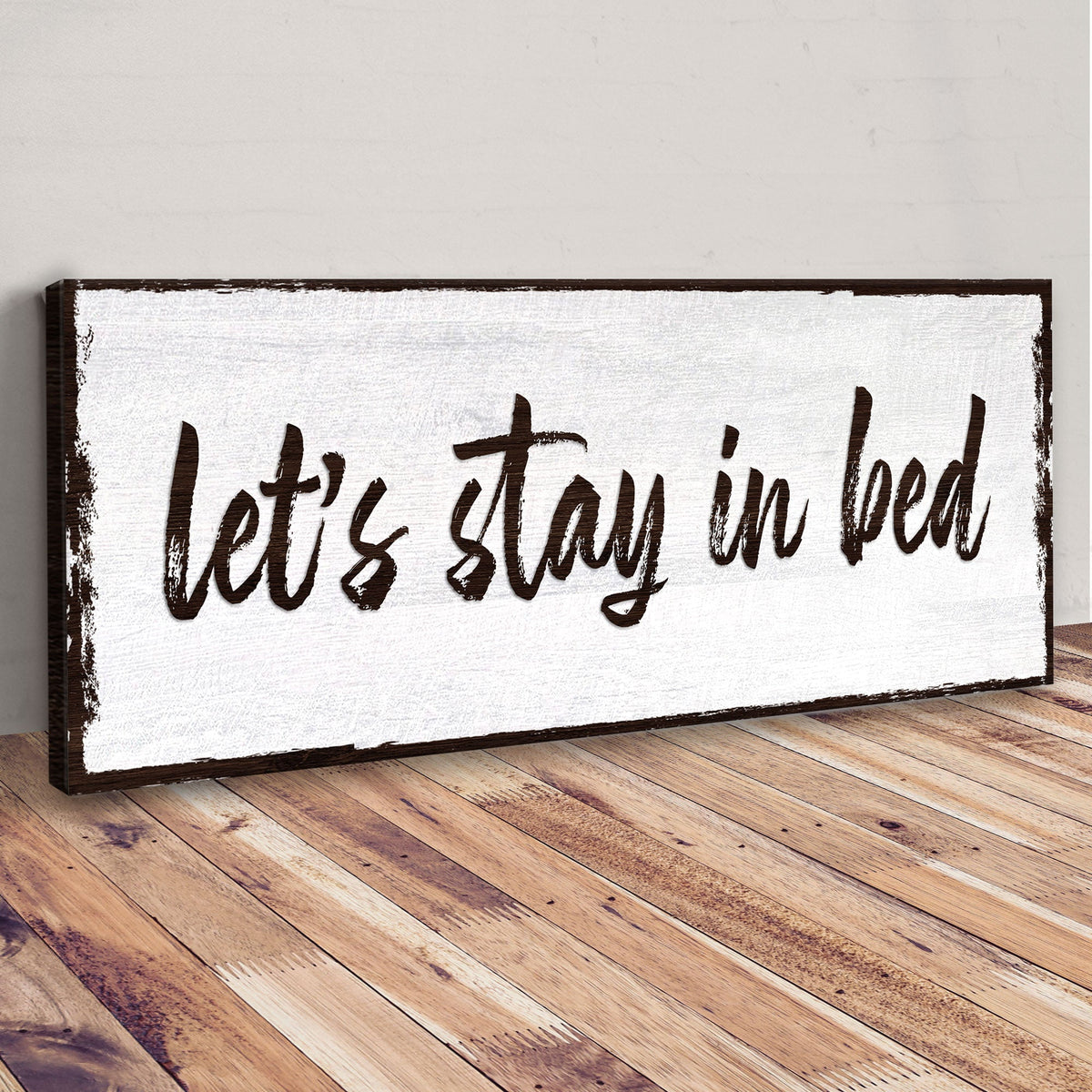 Angled view of the Let's Stay in Bed brush script canvas sign leaning on a wood floor