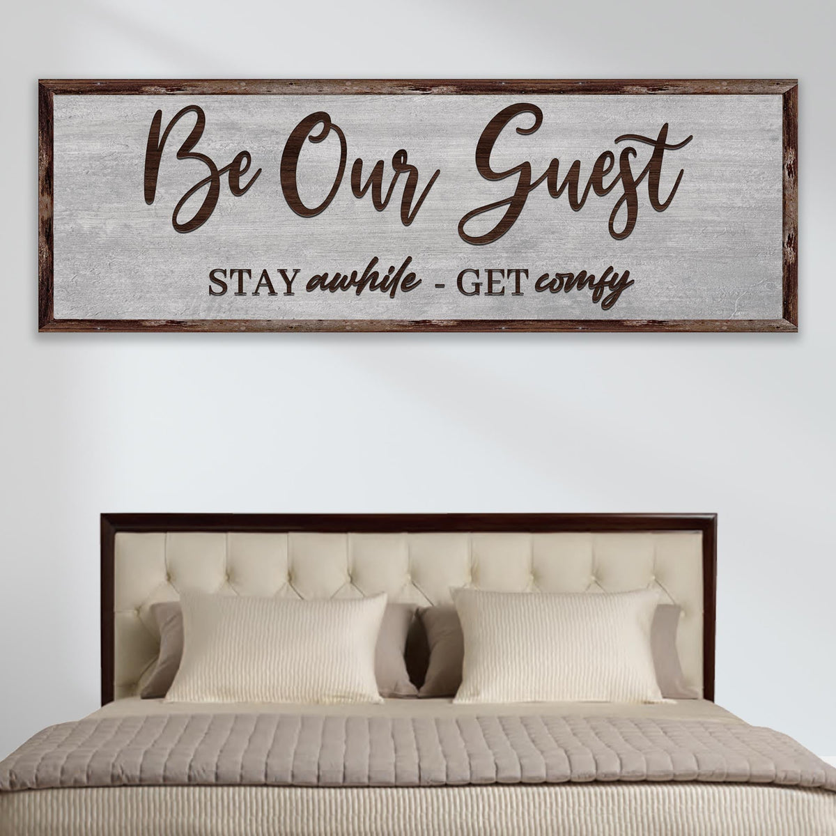Long gray canvas sign reading Be Our Guest in brown brush script with a second line below, above a tufted bed
