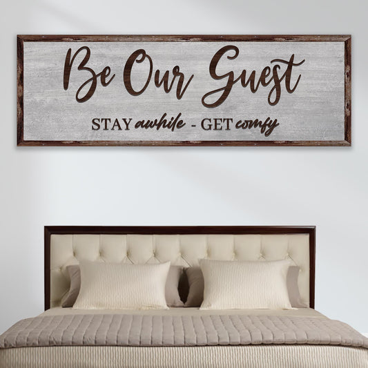 Long gray canvas sign reading Be Our Guest in brown brush script with a second line below, above a tufted bed