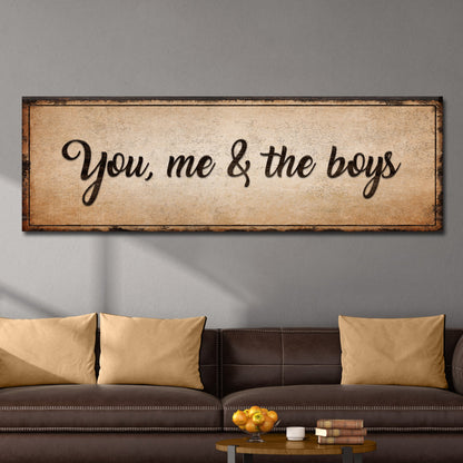 Long tan canvas sign with bold dark script reading You, me & the boys above a dark leather sofa with tan pillows